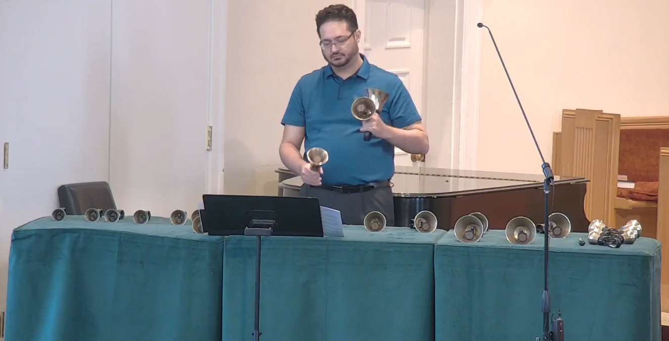 A man stands at a table equipped with handbells, in a room possibly with musical instruments nearby, awaiting an audience.